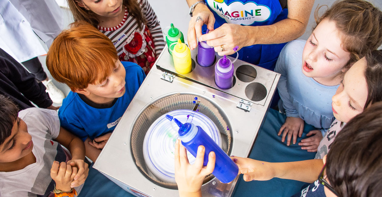 A boy pouring a blue bottle of paint into the spin art machine while the Crayola Imagine Arts Instructor and 5 other children